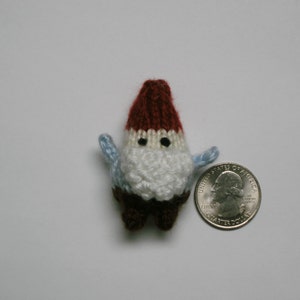 Op de afbeelding: Een kleine gebreide gnome met een rood-witte muts, witte baard en blauwe armen. De gnome is ongeveer zo groot als een kwartje.