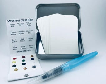 May include: An open, gray metal tin containing a stack of white cards, a color guide with color dot samples, and a blue watercolor brush. The color guide lists paint colors such as Mayan Red, Lemon Yellow, and Black Ochre.