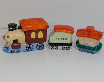 Op de afbeelding: Een keramische zout- en peperstrooier set in de vorm van een trein. De locomotief is beige, bruin en blauw, met een zwarte schoorsteen. De peperstrooier is groen met "PEPPER" erop gedrukt. De zoutstrooier is wit met "SALT" erop gedrukt.
