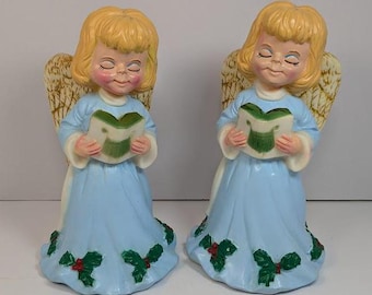 Op de afbeelding: Twee blauwe en witte engelbeeldjes met blond haar en gesloten ogen. Elke engel houdt een boek vast en draagt een rand van hulstbladeren.