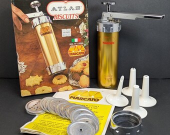 Op de afbeelding: Een goudkleurige Marcato koekjespers met een zilveren handvat en diverse koekjesvormschijven. De doos leest "Atlas Biscuits" en toont koekjes. Inclusief witte plastic spuitmonden en een metalen ring.