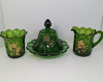 Op de afbeelding: Een groene glazen serveerset met een botervloot met deksel, een melkkan en een tumbler. Elk stuk is versierd met gouden bloemmotieven en geschulpte randen.