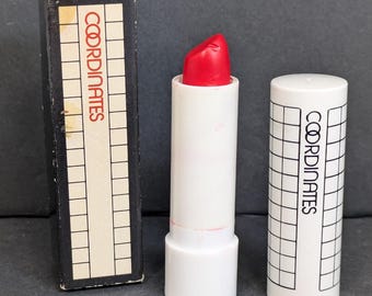 Op de afbeelding: Een rode lipstick met een witte tube en dop, naast een zwarte doos. De lipstick is voorzien van het opschrift "COORDINATES" in rode letters. De tube en dop hebben een rasterpatroon.