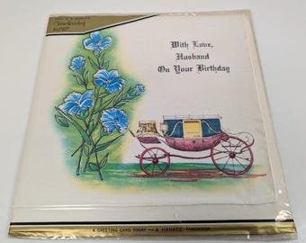 Op de afbeelding: Een witte kaart met een blauw en groen bloemenontwerp en een illustratie van een koets. De kaart heeft de tekst "With Love, Husband On Your Birthday".