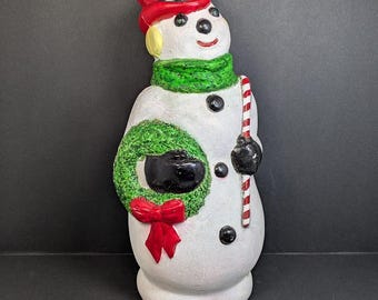 Op de afbeelding: Een wit sneeuwpopfiguur met een rode hoge hoed en een groene sjaal. De sneeuwpop houdt een groene krans met een rode strik in de ene hand en een rood-wit gestreepte zuurstok in de andere hand. Het heeft zwarte knopen en ogen.