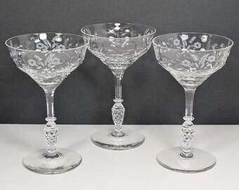 Op de afbeelding: Drie heldere champagne coupe glazen. Elk glas heeft een brede kom met bloemetsing, een diamantgeslepen steel en een ronde voet. De glazen zijn gerangschikt op een wit oppervlak tegen een donkergrijze achtergrond.