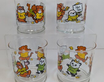 Op de afbeelding: Een set van vier doorzichtige glazen met kleurrijke Shirt Tales stripfiguren. De glazen tonen illustraties van Rick, Tyg, Digger en Pammy die shirts dragen met hun namen erop gedrukt. Elk glas heeft een platte bodem.