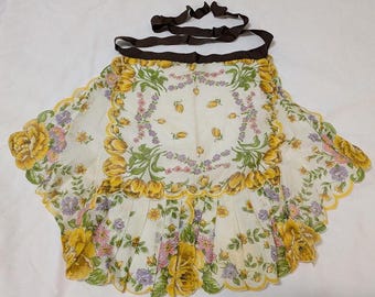 Op de afbeelding: Een witte schort met een bloemenprint met gele en paarse bloemen. De schort heeft een bruin lint om te strikken en een gegolfde rand.