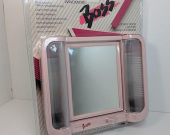 Op de afbeelding: Een verlichte Windmere Boss make-upspiegel in de originele plastic verpakking. De roze rechthoekige spiegel is voorzien van zijdelingse lichtverspreiders, een verstelbare standaard en een aan/uit-schakelaar. De verpakking vermeldt model C606R/576.