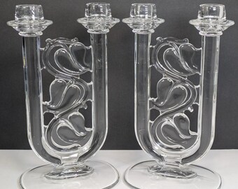 Op de afbeelding: Twee glazen kandelaars. Elke kandelaar heeft een ronde voet, een U-vormige steel met een decoratief bloemmotief en twee kaarshouders. De kandelaars zijn gemaakt van helder glas.