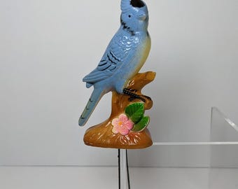 Op de afbeelding: Een blauwe en gele keramische vogelfiguur, gepercht op een bruine tak met een roze bloem. De vogel heeft een zwarte kuif en staat op een bruine basis.
