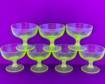 Op de afbeelding: Acht neon groene dessertglazen met steel zijn gerangschikt op een paars oppervlak. De glazen hebben een afgeronde komvorm en een korte steel. De glazen zijn gemaakt van transparant materiaal.