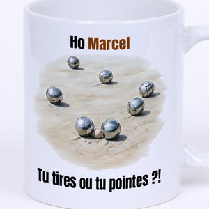 Könnte beinhalten: Weiße Keramik-Tasse mit einer Grafik von Metallkugeln auf Sand. Die Tasse trägt die Aufschrift "Ho Marcel" und "Tu tires ou tu pointes ?!". Die Tasse hat einen gebogenen Griff.