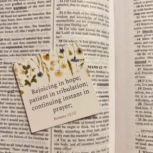May include: A close-up of a Bible verse from Romans 12:12, "Rejoicing in hope; patient in tribulation; continuing instant in prayer." The verse is printed on a white card with a floral design.