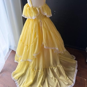 Op de afbeelding: Een felgele, vloerlange jurk met off-shoulder ruche-mouwen, een gelaagde rok en een fijne witte kanten rand langs de mouwen en de zoom. Het lijfje is voorzien van dunne schouderbandjes en heeft een volledig, zwierig silhouet.