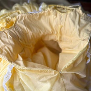 Op de afbeelding: Een lichtgeel kledingstuk met een geplooide halslijn en witte kanten afwerking langs de randen. Het item is voorzien van een zichtbare ritssluiting aan de zijkant en een lichte, soepele stof met diverse plooien en geplooide secties.