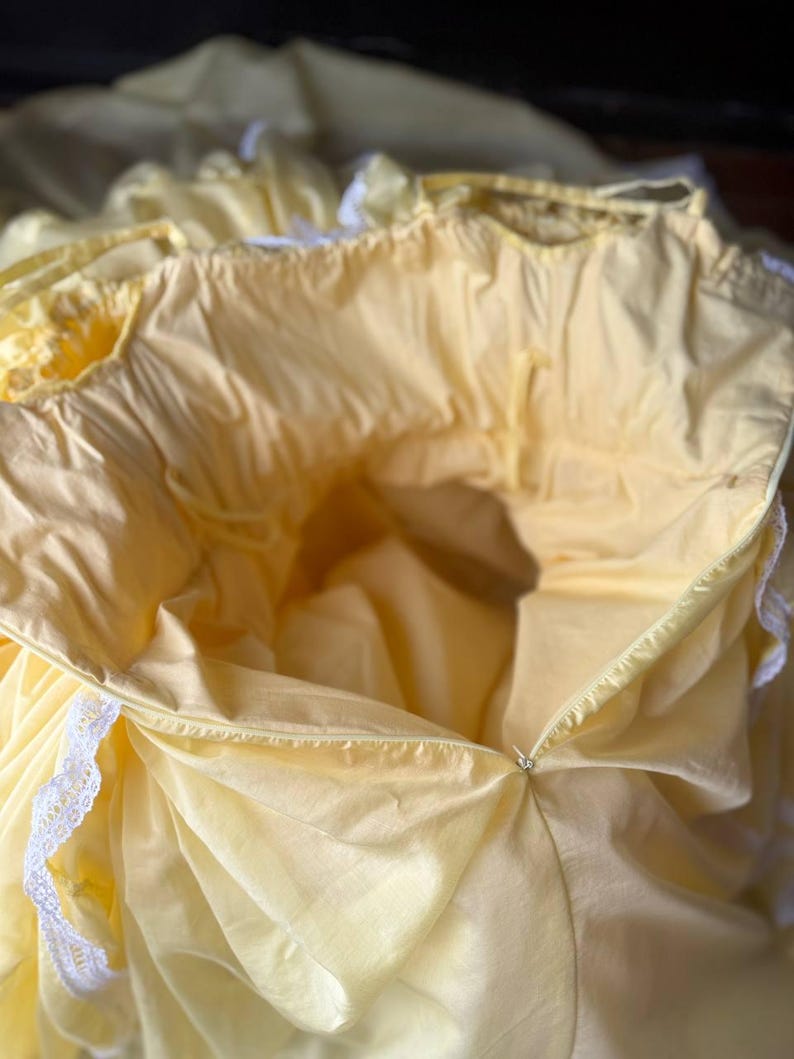 Op de afbeelding: Een lichtgeel kledingstuk met een geplooide halslijn en witte kanten afwerking langs de randen. Het item is voorzien van een zichtbare ritssluiting aan de zijkant en een lichte, soepele stof met diverse plooien en geplooide secties.