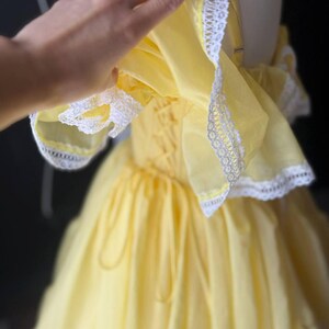 Op de afbeelding: Een felgele jurk met off-shoulder ruchemouwen afgezet met witte kant. Het lijfje is voorzien van een vetersluiting aan de zijkant met bijpassend geel lint, en de rok heeft een vol, geplooid silhouet met meerdere lagen stof.