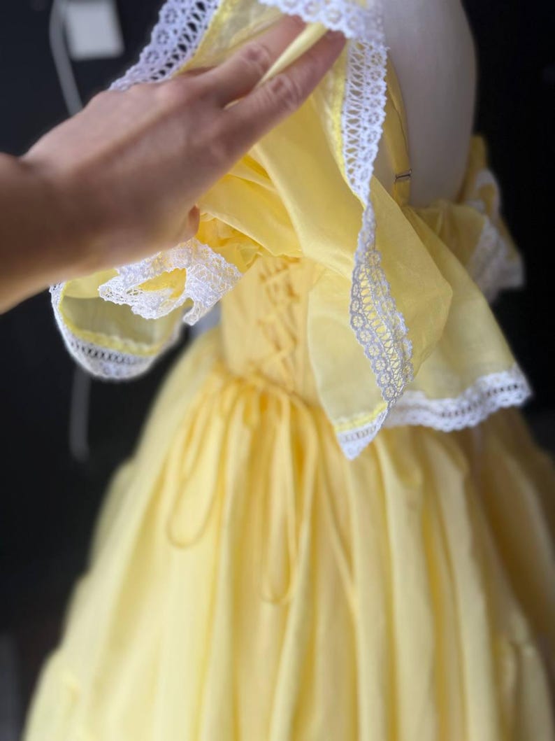 Op de afbeelding: Een felgele jurk met off-shoulder ruchemouwen afgezet met witte kant. Het lijfje is voorzien van een vetersluiting aan de zijkant met bijpassend geel lint, en de rok heeft een vol, geplooid silhouet met meerdere lagen stof.