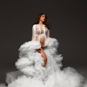 May include: A white, layered tulle maternity gown with a sheer robe. The gown features a flowing skirt and a delicate, open-front robe. The model is wearing a bra and a necklace. The background is a neutral gray.