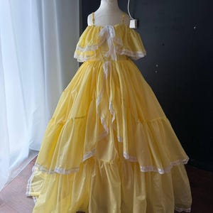 Op de afbeelding: Een vloerlange gele jurk met een off-shoulder halslijn met ruches, dunne schouderbandjes en witte kanten afwerking langs de randen van de ruches en de zoom. De rok is wijd en gelaagd met een volumineus silhouet.