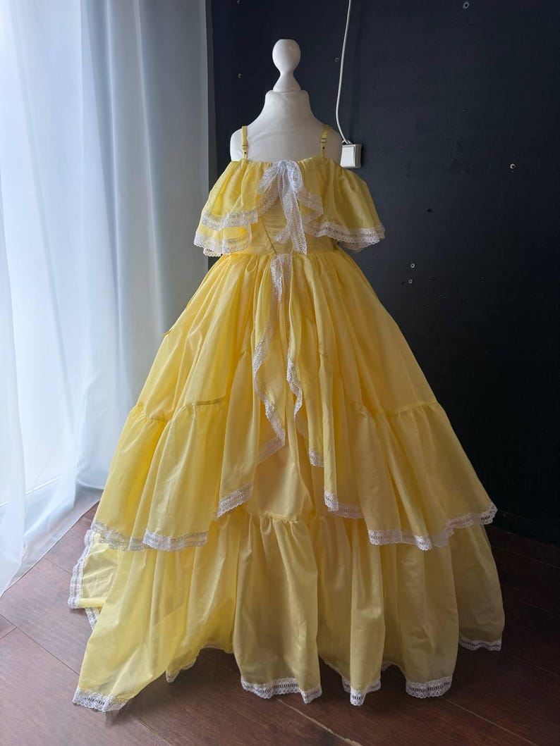 Op de afbeelding: Een vloerlange gele jurk met een off-shoulder halslijn met ruches, dunne schouderbandjes en witte kanten afwerking langs de randen van de ruches en de zoom. De rok is wijd en gelaagd met een volumineus silhouet.