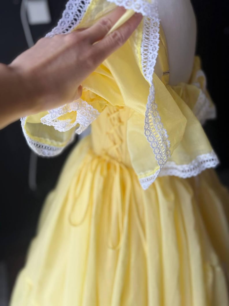 Op de afbeelding: Een felgele jurk met off-shoulder ruchemouwen afgezet met witte kant. Het lijfje is voorzien van een vetersluiting aan de zijkant met bijpassend geel lint, en de rok heeft een vol, geplooid silhouet met meerdere lagen stof.