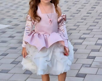 May include: A young girl in a light pink and white dress with long sleeves and a layered skirt. The dress has a square neckline and a pink bow on the sleeve. She wears a pendant necklace and beige sandals.