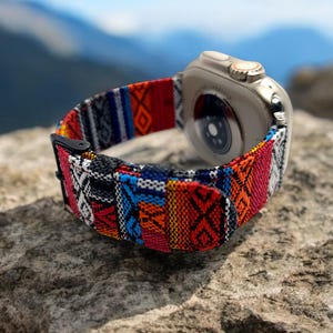 Może przedstawiać: Smartwatch z kolorowym, tkanym paskiem z geometrycznymi wzorami w kolorach czerwonym, niebieskim, pomarańczowym i czarnym. Srebrny zegarek spoczywa na teksturowanej skale, z rozmytym tłem gór.