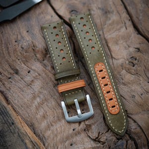 May include: Olive green leather watch straps with perforated holes and orange detailing. The straps have white stitching and a silver buckle. The watch bands are shown on a wooden surface.