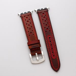 May include: A brown leather watch band with red stitching and perforated design. The band is next to a silver Apple Watch on a wooden surface. The text "No:5" is visible in the lower left corner.