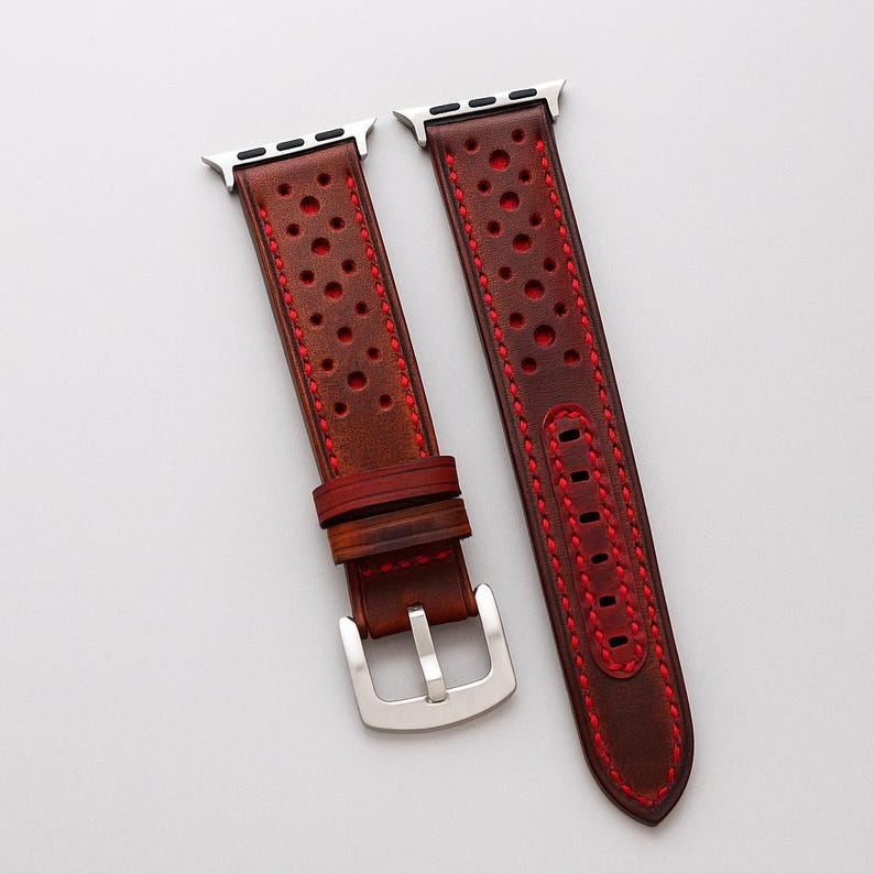 May include: A brown leather watch band with red stitching and perforated design. The band is next to a silver Apple Watch on a wooden surface. The text "No:5" is visible in the lower left corner.