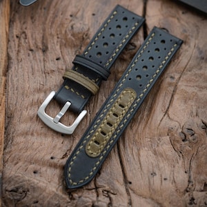 May include: Black leather watch strap with olive green detailing. The strap is perforated with small holes and has olive green stitching. A silver-coloured buckle is attached. The strap is made of textured leather.