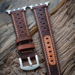 May include: Two brown leather watch straps with silver clasps and connectors. The straps feature a perforated design and are stitched with white thread. The leather has a distressed look, displayed on a wooden surface.