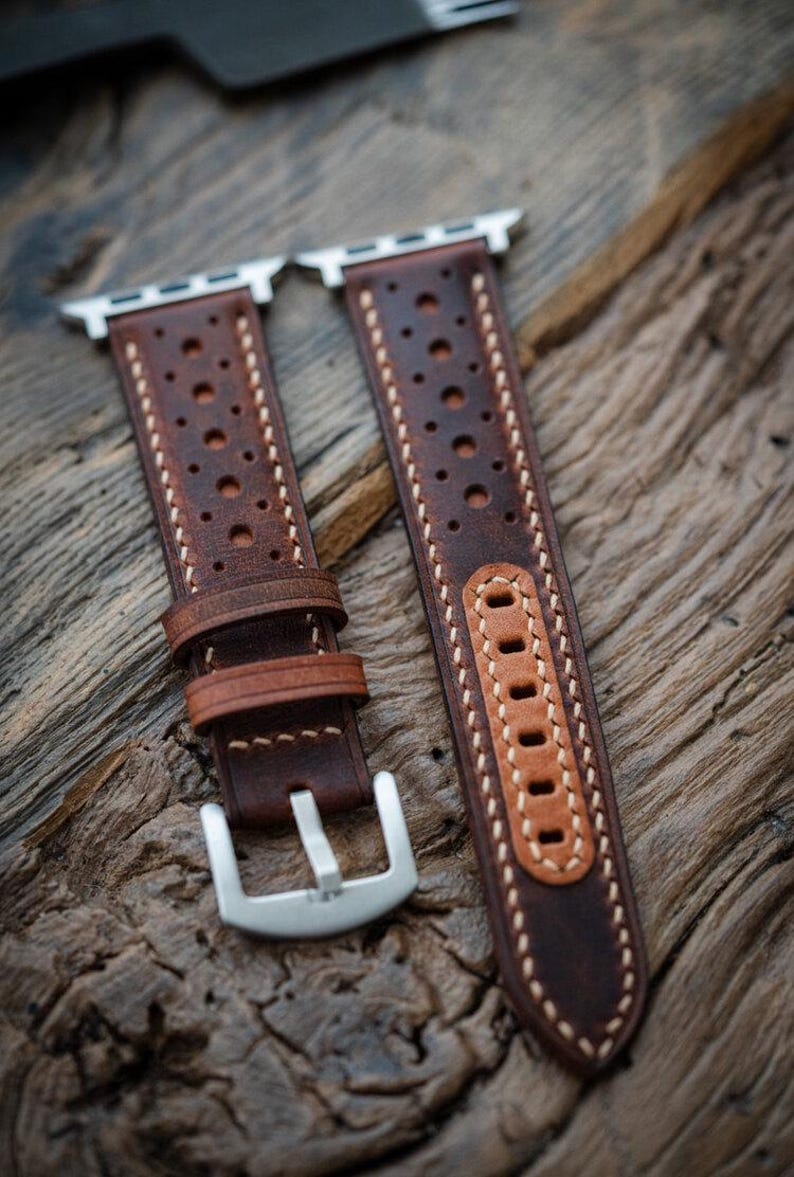 May include: Two brown leather watch straps with silver clasps and connectors. The straps feature a perforated design and are stitched with white thread. The leather has a distressed look, displayed on a wooden surface.