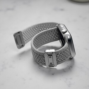 May include: A silver smartwatch featuring a light gray woven nylon sport loop band. The band includes a metallic rectangular adjustment buckle with two visible screws and a matching silver end tab. The watch case has a side crown and button.