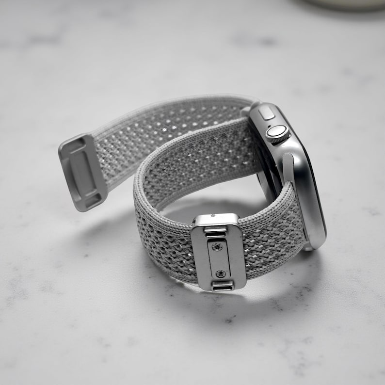 May include: A silver smartwatch featuring a light gray woven nylon sport loop band. The band includes a metallic rectangular adjustment buckle with two visible screws and a matching silver end tab. The watch case has a side crown and button.