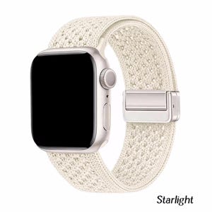 May include: A smartwatch with a rectangular silver case and a black screen, featuring a textured, off-white woven fabric band. The band includes a metallic silver clasp mechanism. The word Starlight is printed in black text below the watch.