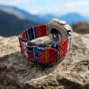 May include: A smartwatch with a colorful woven band featuring geometric patterns in red, blue, orange, and black. The watch is silver and is resting on a textured rock, with a blurred mountain backdrop.
