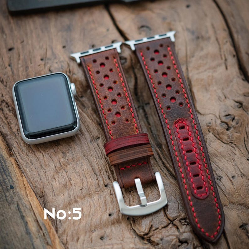 May include: A brown leather watch strap with red stitching and a perforated pattern. A silver Apple Watch sits beside the strap on a wooden surface. The text "No:5" is visible in the lower left corner.
