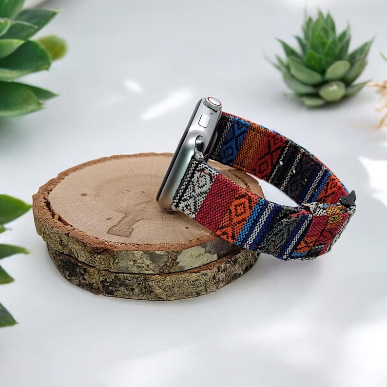 May include: A silver smartwatch with a colorful woven band featuring red, orange, blue, and black geometric patterns. The watch rests on a round, natural wood slice, showcasing a rustic aesthetic.