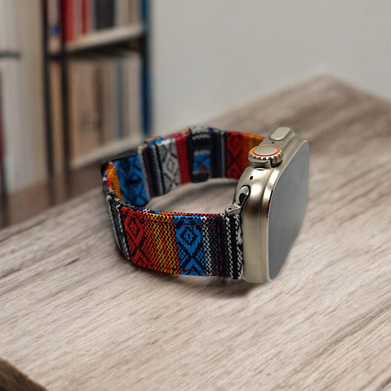 May include: A silver smartwatch with a colorful patterned band. The band features a geometric design with red, blue, yellow, and black accents. The watch is resting on a wooden surface.