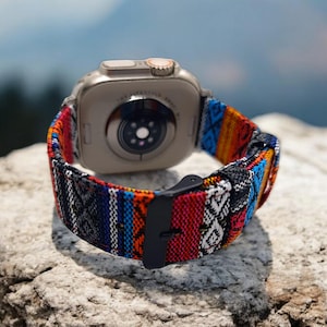 May include: A silver smartwatch with a colorful woven band. The band features a pattern of red, blue, orange, and white stripes and geometric designs. The watch is resting on a textured rock, with a blurred mountain backdrop.