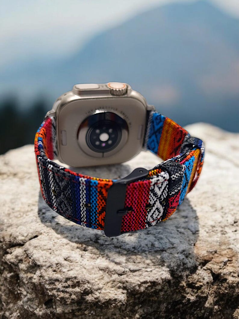 May include: A silver smartwatch with a colorful woven band. The band features a pattern of red, blue, orange, and white stripes and geometric designs. The watch is resting on a textured rock, with a blurred mountain backdrop.