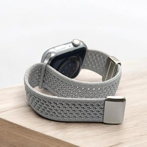 May include: A silver smartwatch featuring a light gray, textured, woven elastic watch band. The band includes metallic silver end connectors and a matching silver buckle mechanism, showcasing a flexible, breathable, and lightweight design.