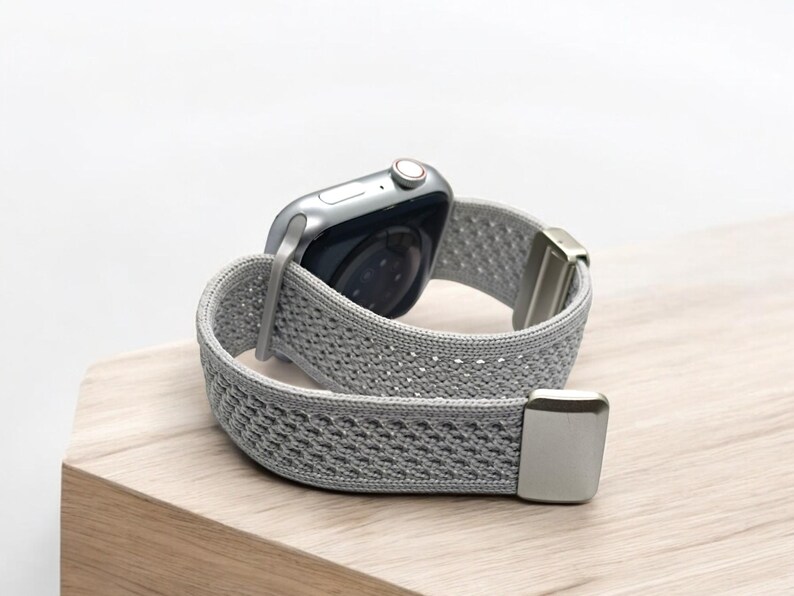 May include: A silver smartwatch featuring a light gray, textured, woven elastic watch band. The band includes metallic silver end connectors and a matching silver buckle mechanism, showcasing a flexible, breathable, and lightweight design.