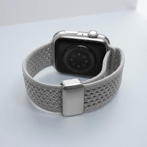 May include: A silver smartwatch featuring a light gray, textured, woven elastic watch band. The watch case has a metallic finish with a side crown and button, while the band includes a rectangular silver-toned metal sliding adjustment buckle.