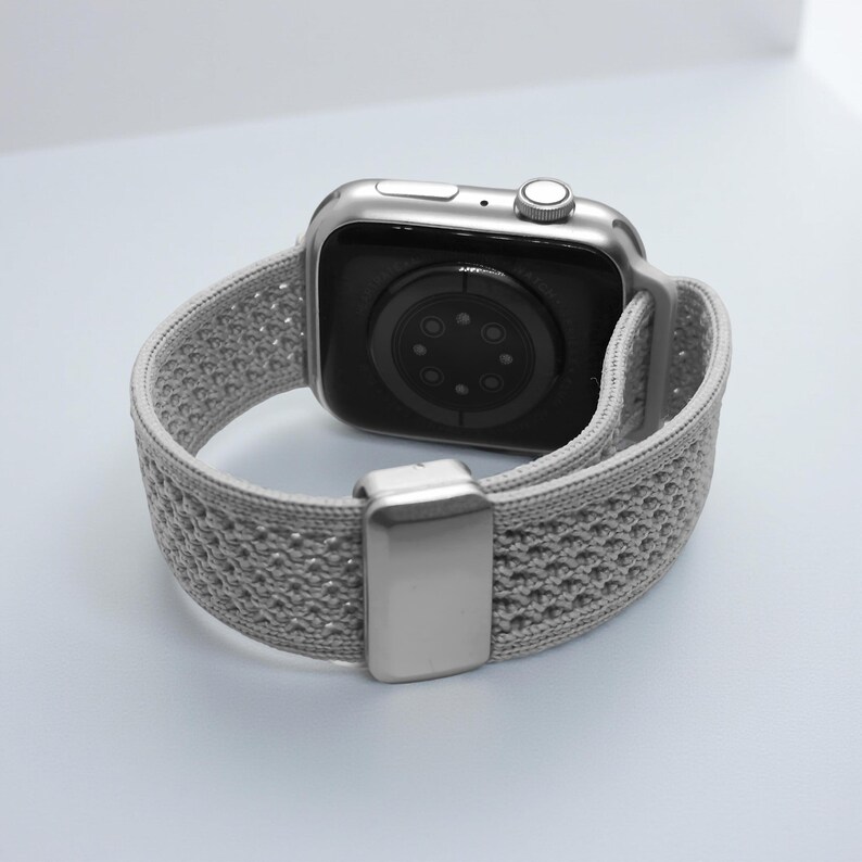 May include: A silver smartwatch featuring a light gray, textured, woven elastic watch band. The watch case has a metallic finish with a side crown and button, while the band includes a rectangular silver-toned metal sliding adjustment buckle.