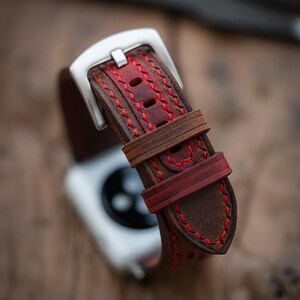 May include: Close-up of a brown leather watch strap with red stitching. The strap has a silver buckle and a contrasting red accent. The watch strap is designed for a smartwatch.