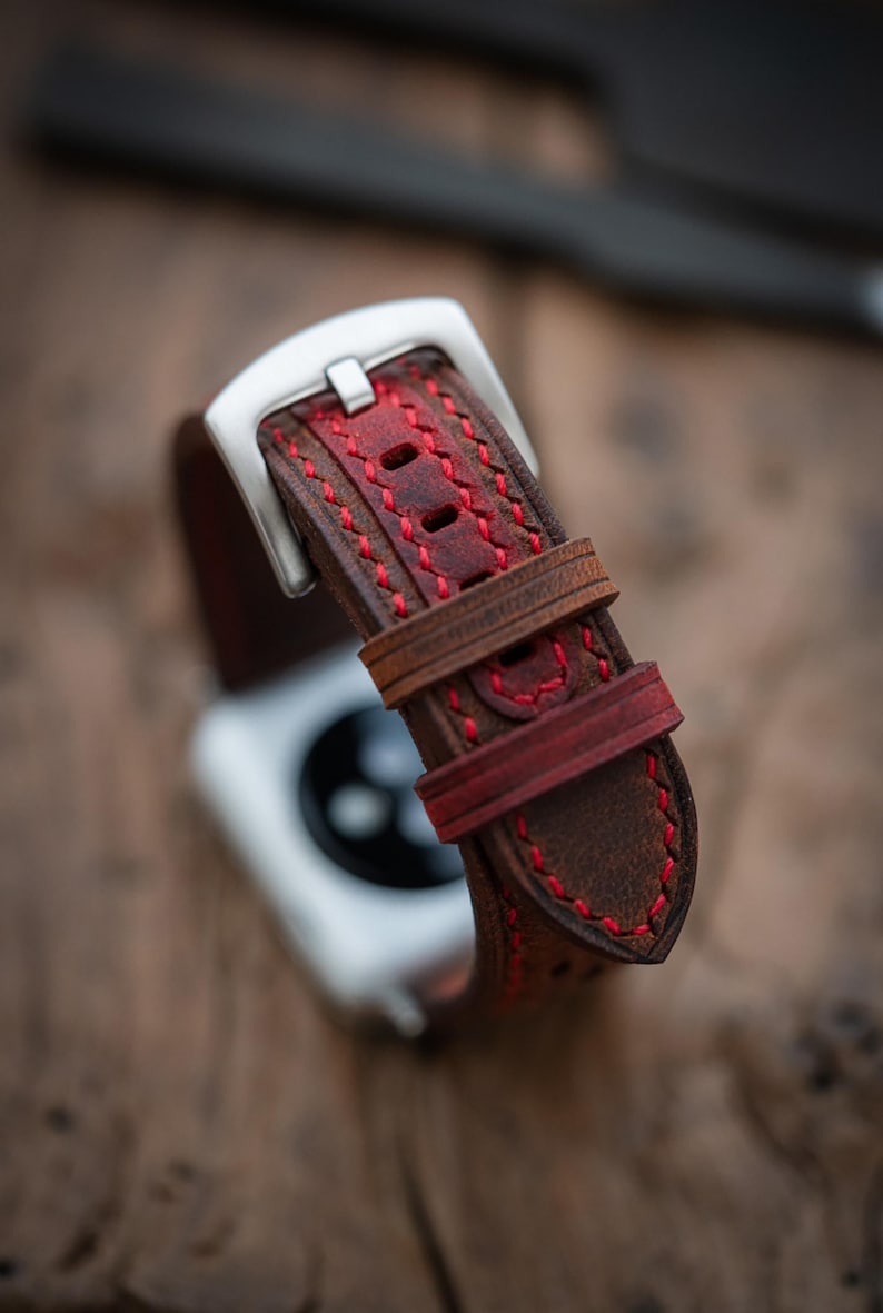 May include: Close-up of a brown leather watch strap with red stitching. The strap has a silver buckle and a contrasting red accent. The watch strap is designed for a smartwatch.